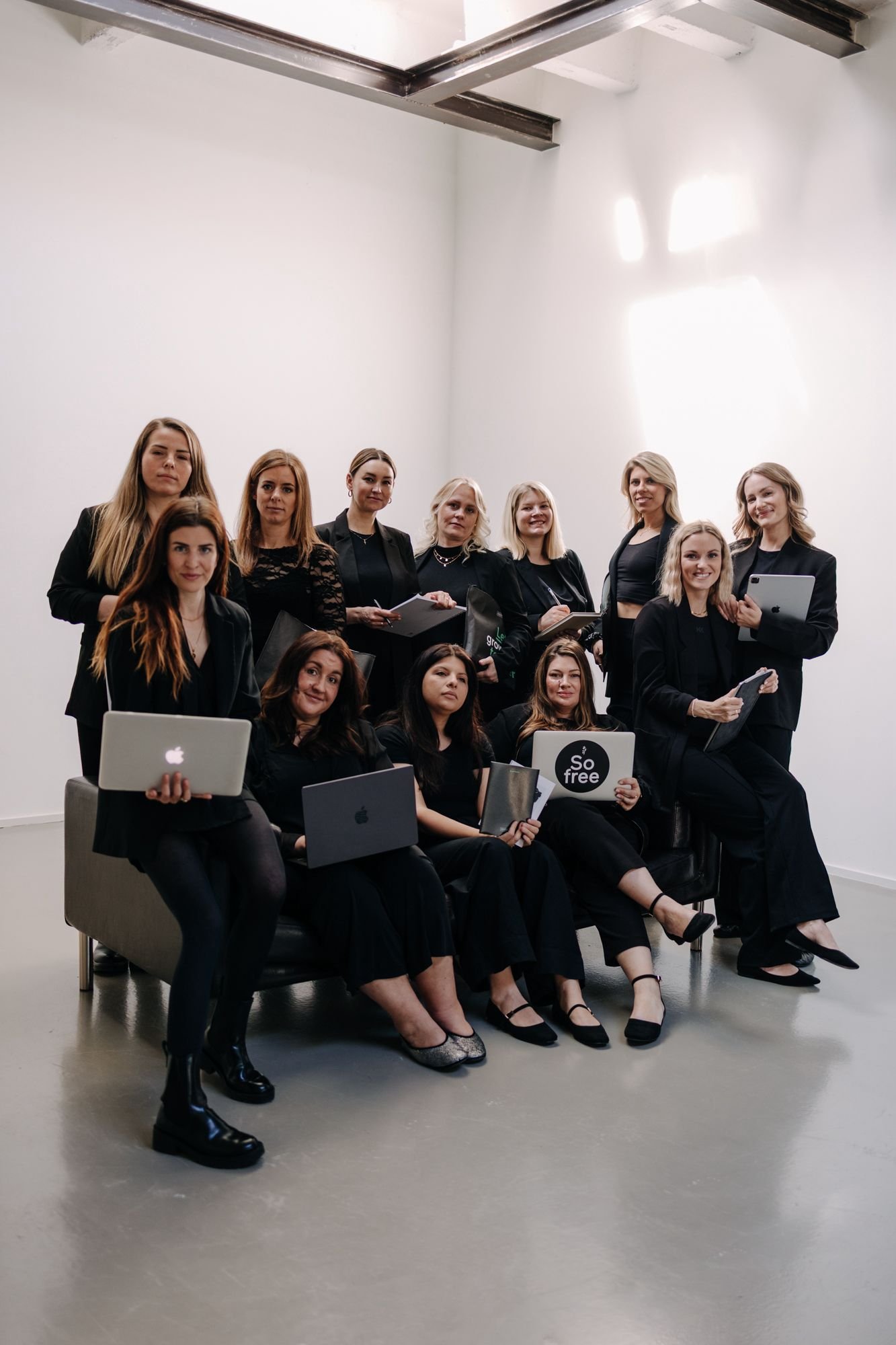 Dreizehn Frauen in schwarzer Kleidung in einem hellen Studio, einige sitzend, einige stehend